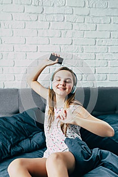 Beautiful young Caucasian teenage girl sitting in bed with modern headphones is holding phone in her hand, dancing to cool music a