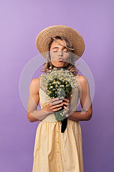 Beautiful young caucasian woman closes eyes, compresses lips, holds daisies on purple background.