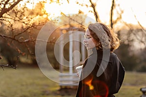 Beautiful young Caucasian woman enjoy sunny spring day, park cherry garden, romantic portrait, drinking coffee, lady in black coat