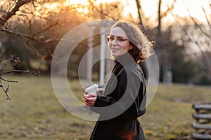 Beautiful young Caucasian woman enjoy sunny spring day, park cherry garden, romantic portrait, drinking coffee, lady in black coat