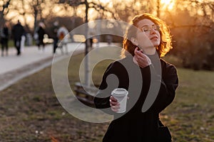 Beautiful young Caucasian woman enjoy sunny spring day, park cherry garden, romantic portrait, drinking coffee, lady in black coat
