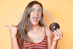 Beautiful young caucasian woman holding donut pointing thumb up to the side smiling happy with open mouth