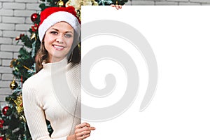 Beautiful and young caucasian woman holding and pointing to blank white board in front of big christmas tree. Concept for