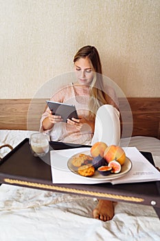 Beautiful young caucasian woman sitting on bed with sleepy look in morning. Working on bed remotely, working from home, busy