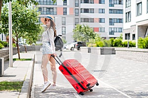 Beautiful young caucasian woman in a summer hat posing with a red suitcase outdoors. Happy smiling girl goes on vacation