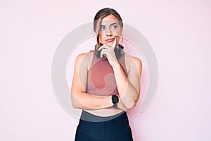 Beautiful young caucasian woman wearing gym clothes and using headphones serious face thinking about question with hand on chin,