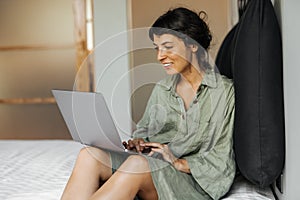 Beautiful young caucasian woman working at her laptop on white bed in room.