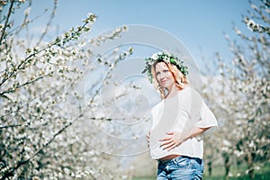 Beautiful young cheerful pregnant woman in wreath of flowers on head touching belly while walking in spring tree garden. Beauty