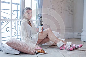 Beautiful young cheerful winter woman sitting on floor and drinking coffee indoor