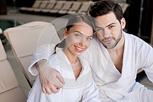 beautiful young couple in bathrobes embracing and smiling at camera in spa