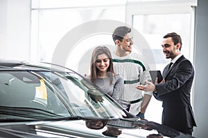 Beautiful young couple choosing a car at the dealership talking to the salon manager with tablet in hands