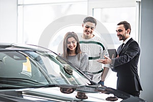 Beautiful young couple choosing a car at the dealership talking to the salon manager with tablet in hands