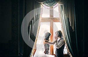 beautiful young couple, costs near a window in an ancient interior, the groom with tenderness and love kisses the bride& x27;s