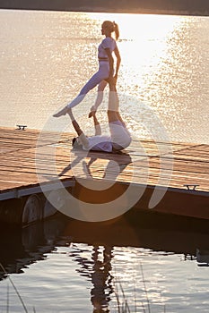 Beautiful young couple doing acro yoga on the pier against sunset. Man lying on the pier and balancing woman in his feet
