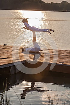 Beautiful young couple doing acro yoga on the pier against sunset. Man lying on the pier and balancing woman in his feet