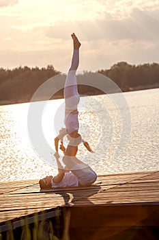 Beautiful young couple doing acro yoga on the pier against sunset. Man lying on the pier and balancing woman in his feet