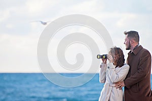 Happy beautiful young couple fashionable man and woman in autumn coat looking through binoculars near sea with horizon and flying