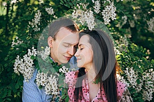 Beautiful young couple gently hugging in green leaves and white flowers in spring garden in sunshine. Happy family embracing at