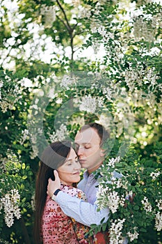 Beautiful young couple gently hugging in green leaves and white flowers in spring garden in sunshine. Happy family embracing at