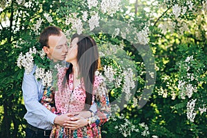 Beautiful young couple gently hugging and kissing at green leaves and white flowers in spring garden. Happy family embracing at