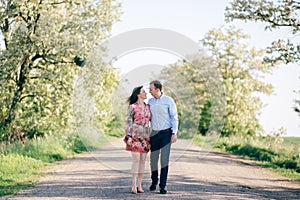 Beautiful young couple holding hands and walking on road in sunshine among spring field and trees. Happy family in love relaxing