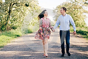 Beautiful young couple holding hands and walking on road in sunshine among spring field and trees. Happy family in love relaxing