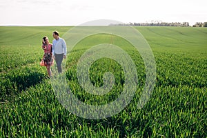 Beautiful young couple holding hands and walking in sunshine in spring meadow. Happy family embracing in green field in sunlight.