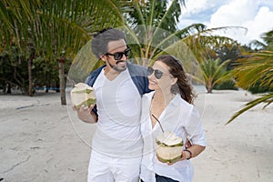Beautiful young couple joyfully on a tropical beach with coconuts in their hands on the seashore under a green palm tree.
