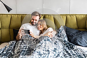 Beautiful young couple in love at home laying in bed, using mobile phone