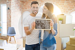 Beautiful young couple moving to a new house, smiling very happy holding blackboard with our first home text