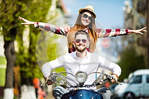 Beautiful young couple riding scooter together while happy woman raising arms and smiling