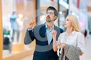 Beautiful young couple searching store in shopping mall using mobile phone, looking around and pointing fingers.