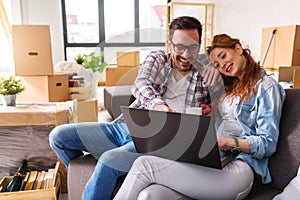 Beautiful young couple shopping on-line using a laptop computer and smiling while sitting in their new apartment after move