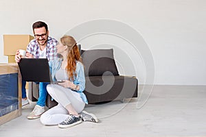 Beautiful young couple shopping on-line using a laptop computer and smiling while sitting in their new apartment after move