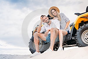 beautiful young couple sitting on ATV on sandy dune and looking