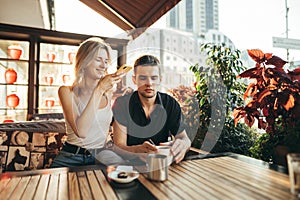 Beautiful young couple sitting on terrace at restaurant at sunset and drinking coffee, happy girl taking photo on smartphone