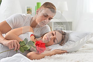 Beautiful young couple on soft bed in morning. Man giving woman a flower