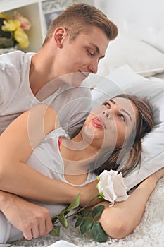 Beautiful young couple on soft bed in morning. Man giving woman a flower