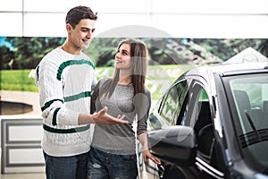 Beautiful young couple standing at the dealership choosing the car to buy. Man pointed on car.