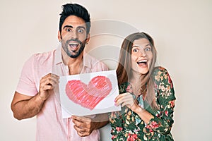 Beautiful young couple together holding heart draw celebrating crazy and amazed for success with open eyes screaming excited