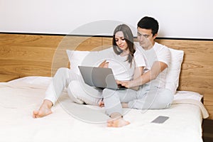 Beautiful young couple is using laptop and smiling while lying in bed at home