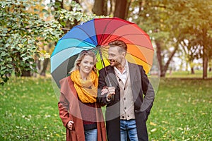 A beautiful young couple is walking in the park under a rainbow colored umbrella. A beautiful girl walks through the