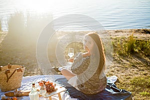 Beautiful young curly woman in warm cardigan on cozy autumn picnic by the lake with fruits, pastries and glass of white wine. Sun