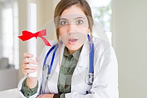 Beautiful young doctor woman holding degree scared in shock with a surprise face, afraid and excited with fear expression