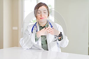 Beautiful young doctor woman wearing medical coat and stethoscope smiling with hands on chest with closed eyes and grateful