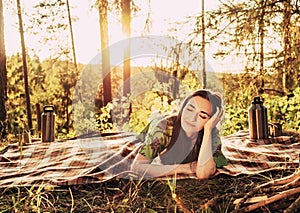 Beautiful young dreaming girl lying on a plaid in a forest glade during sunset bright sunlight around beauty of nature