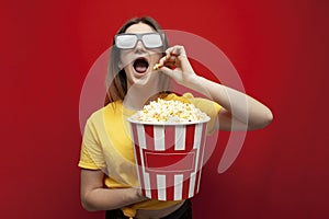 Beautiful young emotional girl in 3D glasses eats popcorn, watches a movie and screams on a red color background, surprised viewer