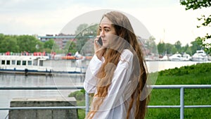 Beautiful young European girl calls and talks by phone near the river on the waterfront, slow motion. Woman with long