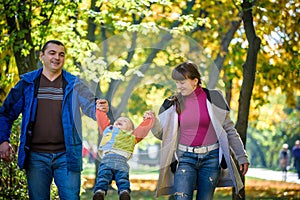 Beautiful young family on a walk in autumn forest on maple yellow trees background. Father and mother hold son on hands. Happy