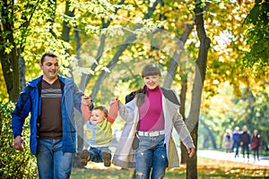 Beautiful young family on a walk in autumn forest on maple yellow trees background. Father and mother hold son on hands. Happy
